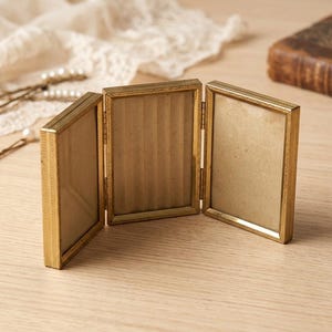 May include: A triptych photo frame featuring three rectangular panels connected by metal hinges. The frame has a metallic gold finish with a textured, etched border pattern around each glass pane. The panels are arranged in a standing, open position.