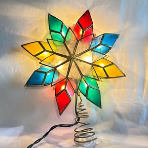 Puede incluir: Una estrella decorativa para la punta del árbol compuesta por paneles de vidrio tintado en rojo, verde, azul y amarillo. La estrella tiene un marco de metal, está iluminada por luces internas y cuenta con una base metálica en espiral.