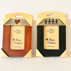 Puede incluir: Dos mini marcos de fotos en embalaje. Un marco es marrón con un corazón recortado, el otro es negro con figuras de palitos. Cada marco mide 5 x 8 cm.