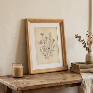 May include: A framed art print featuring a black ink botanical illustration of wildflowers on aged, cream-colored paper. The artwork is encased in a light-toned wooden frame with a white mat, displaying a detailed arrangement of various floral stems.
