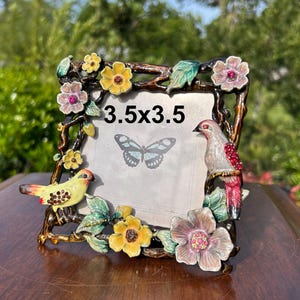 May include: Ornate metal picture frame with a 3.5 x 3.5 inch opening. The frame is decorated with colorful flowers, leaves, and two birds. The frame has a brown finish with yellow, pink, and green accents. A butterfly graphic is visible inside the frame.