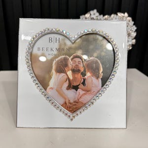 May include: White square photo frame with a heart-shaped opening. The heart is outlined with small, sparkling rhinestones. The frame holds a photo of a man being kissed by two young girls. The text "BH Beekman Home" is visible.