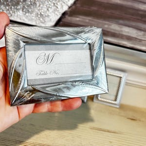 Puede incluir: Un portatarjetas rectangular plateado con un diseño decorativo de hojas. La tarjeta de lugar tiene la letra "M" y las palabras "Table No." impresas. El soporte está en un envoltorio de plástico transparente.