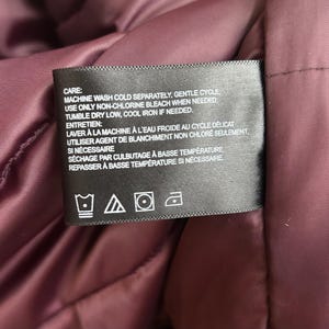 Puede incluir: Una etiqueta negra con texto blanco, cosida en una tela burdeos. La etiqueta proporciona instrucciones de lavado, incluyendo lavado a m&aacute;quina en fr&iacute;o, ciclo suave y secado en secadora a baja temperatura. La etiqueta tambi&eacute;n incluye s&iacute;mbolos de cuidado.