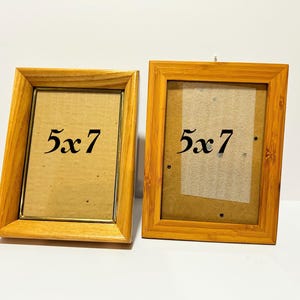 May include: Two wooden picture frames with a light brown finish. Each frame has a 12.7 x 17.8 cm opening and the text "5x7" is printed on the glass. The frames are empty and ready for photos.