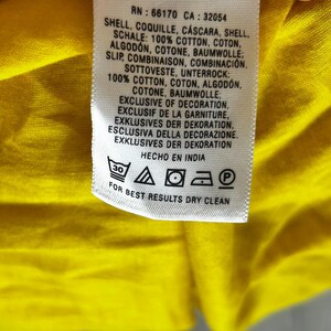 Puede incluir: Primer plano de una etiqueta de ropa con texto en varios idiomas, incluidos ingl&eacute;s, franc&eacute;s y alem&aacute;n. La etiqueta indica que la prenda es 100% algod&oacute;n y proporciona instrucciones de cuidado, incluida la limpieza en seco. La etiqueta est&aacute; sobre una tela amarilla.