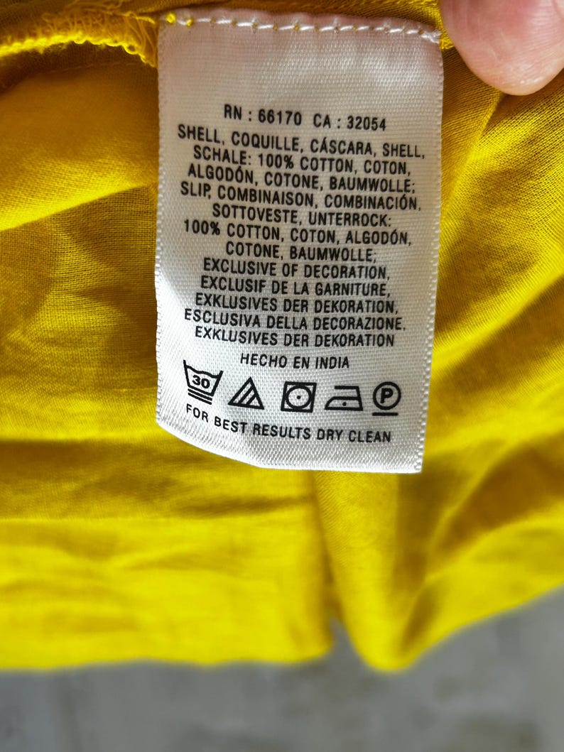Puede incluir: Primer plano de una etiqueta de ropa con texto en varios idiomas, incluidos ingl&eacute;s, franc&eacute;s y alem&aacute;n. La etiqueta indica que la prenda es 100% algod&oacute;n y proporciona instrucciones de cuidado, incluida la limpieza en seco. La etiqueta est&aacute; sobre una tela amarilla.