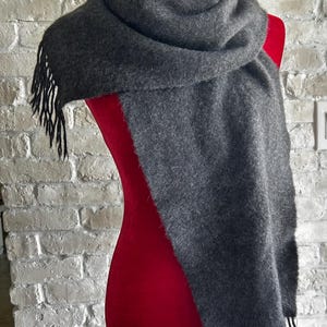 May include: A dark gray scarf with black fringe is draped over a red mannequin. The scarf is made of a soft, textured material. The background is a white brick wall.
