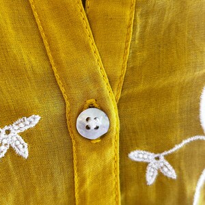 Puede incluir: Primer plano de una blusa amarillo mostaza con cuello en V y cierre de un solo bot&oacute;n. La blusa presenta detalles florales bordados en blanco y una tela delicada y transparente. El bot&oacute;n es de forma redonda y nacarada.