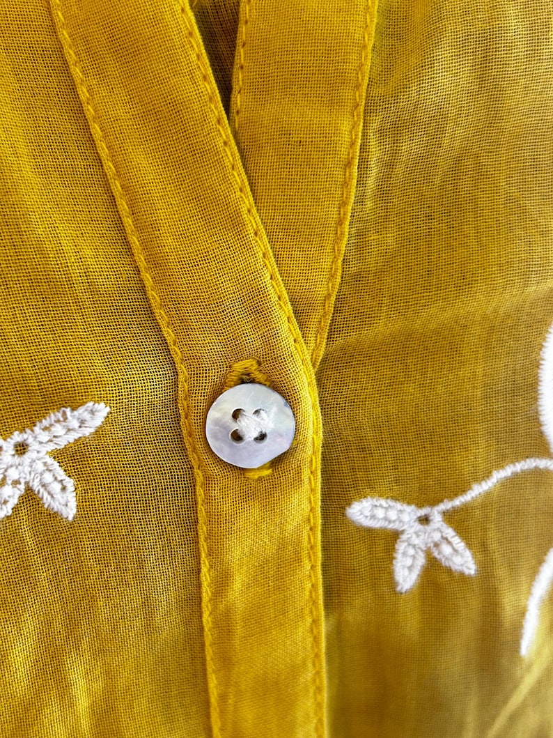 Puede incluir: Primer plano de una blusa amarillo mostaza con cuello en V y cierre de un solo bot&oacute;n. La blusa presenta detalles florales bordados en blanco y una tela delicada y transparente. El bot&oacute;n es de forma redonda y nacarada.