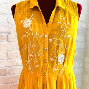 Puede incluir: Vestido sin mangas de color amarillo mostaza con cuello y botones en la parte delantera. El vestido presenta bordados florales blancos y una falda plisada. El vestido se exhibe en un maniqu&iacute; contra una pared de ladrillos blancos.