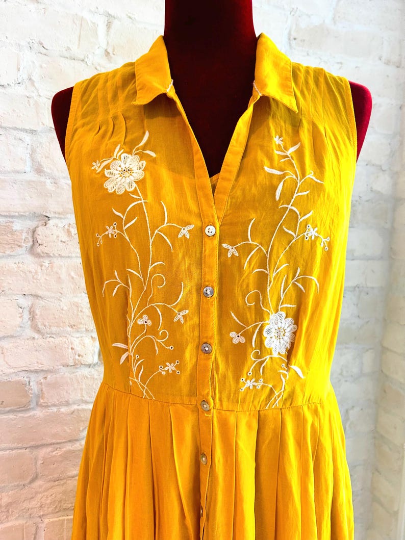 Puede incluir: Vestido sin mangas de color amarillo mostaza con cuello y botones en la parte delantera. El vestido presenta bordados florales blancos y una falda plisada. El vestido se exhibe en un maniqu&iacute; contra una pared de ladrillos blancos.