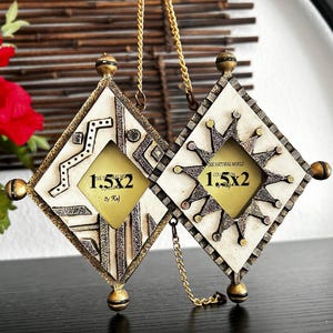 May include: Two diamond-shaped photo frames with gold accents and a cream-colored background. Each frame has a small, gold-colored chain for hanging. The frames have a geometric design and the text "1.5x2" is visible.