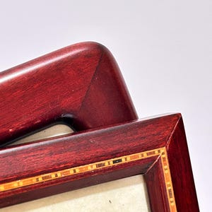 Pode incluir: Uma moldura de madeira de mogno com uma incrustação decorativa de tiras de madeira em tons de marrom, vermelho e dourado. A moldura tem um passe-partout branco e um inserto de papel branco com o texto "Bacon Hill" em escrita cursiva preta.