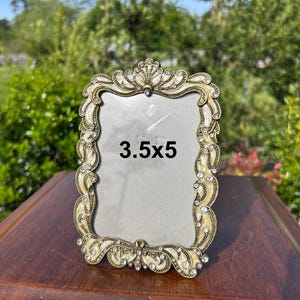 May include: Ornate, antique-style photo frame with a cream and gold finish, embellished with rhinestones. The frame is rectangular and displays the text "3.5x5". The frame is approximately 3.5 inches by 5 inches.