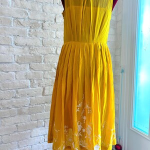 Puede incluir: Vestido sin mangas amarillo mostaza con cuello y falda plisada. El vestido presenta bordados florales blancos cerca del dobladillo. El vestido se exhibe en un maniqu&iacute; contra una pared de ladrillos blancos.
