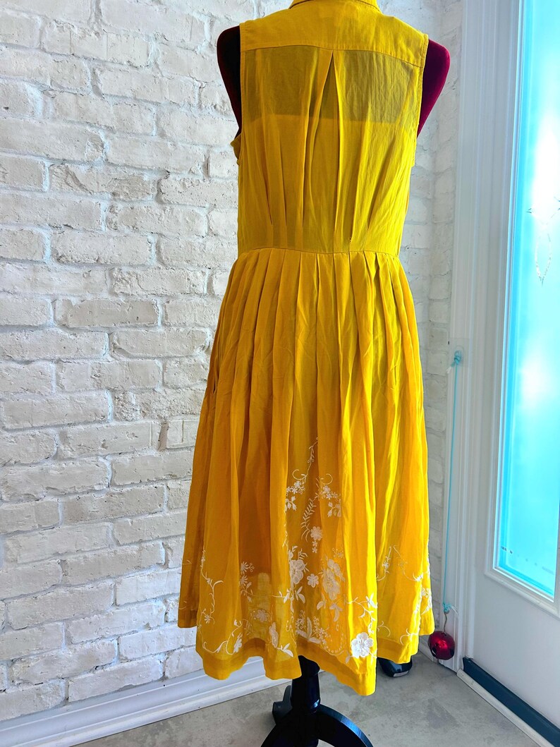 Puede incluir: Vestido sin mangas amarillo mostaza con cuello y falda plisada. El vestido presenta bordados florales blancos cerca del dobladillo. El vestido se exhibe en un maniqu&iacute; contra una pared de ladrillos blancos.