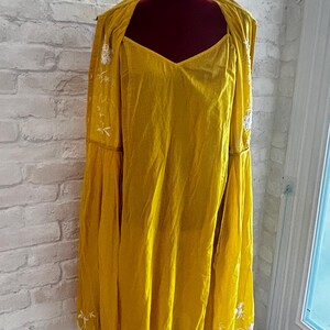 Puede incluir: Conjunto de vestido y chaqueta amarillo mostaza con bordados florales blancos. El vestido tiene cuello en V y la chaqueta tiene mangas largas y acampanadas. El conjunto se exhibe en un maniqu&iacute; negro contra una pared de ladrillos blancos.