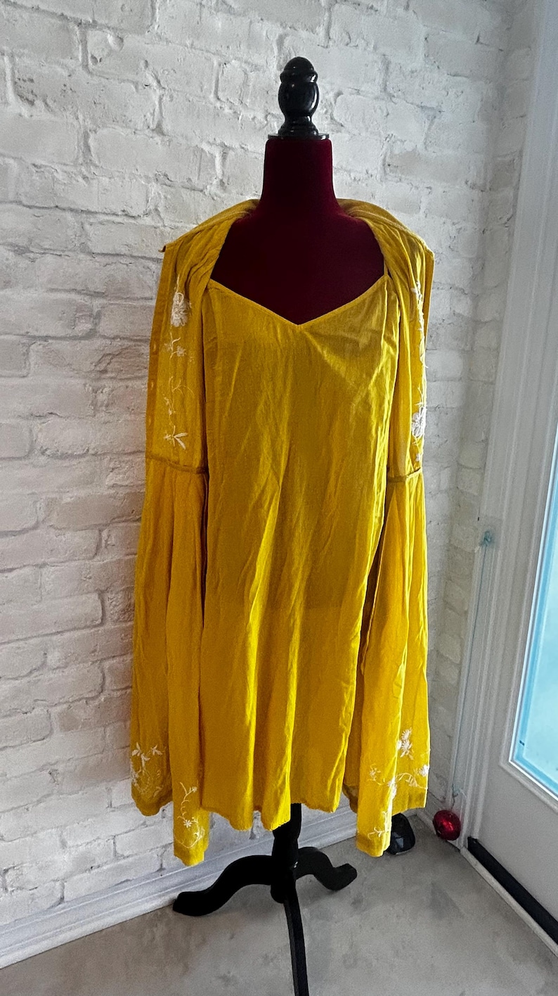 Puede incluir: Conjunto de vestido y chaqueta amarillo mostaza con bordados florales blancos. El vestido tiene cuello en V y la chaqueta tiene mangas largas y acampanadas. El conjunto se exhibe en un maniqu&iacute; negro contra una pared de ladrillos blancos.