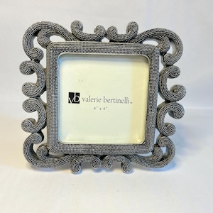 May include: A square picture frame with a textured, metallic silver finish and ornate, swirling scrollwork edges. The center insert features the text "vb valerie bertinelli" and "4" x 4"" printed in black on a white background.