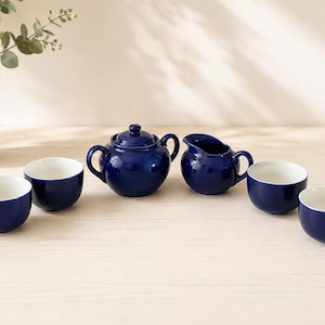 May include: A tea set consisting of a deep blue lidded sugar bowl, a matching creamer pitcher, and four small, round tea cups with white interiors. Each piece features a smooth, glossy finish and a consistent, vibrant cobalt blue exterior color.
