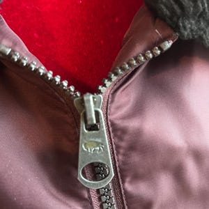 Puede incluir: Primer plano de una cremallera de chaqueta granate. La cremallera plateada tiene un tirador detallado con un dise&ntilde;o de animal. La chaqueta tiene un cuello oscuro y difuso y un interior rojo.