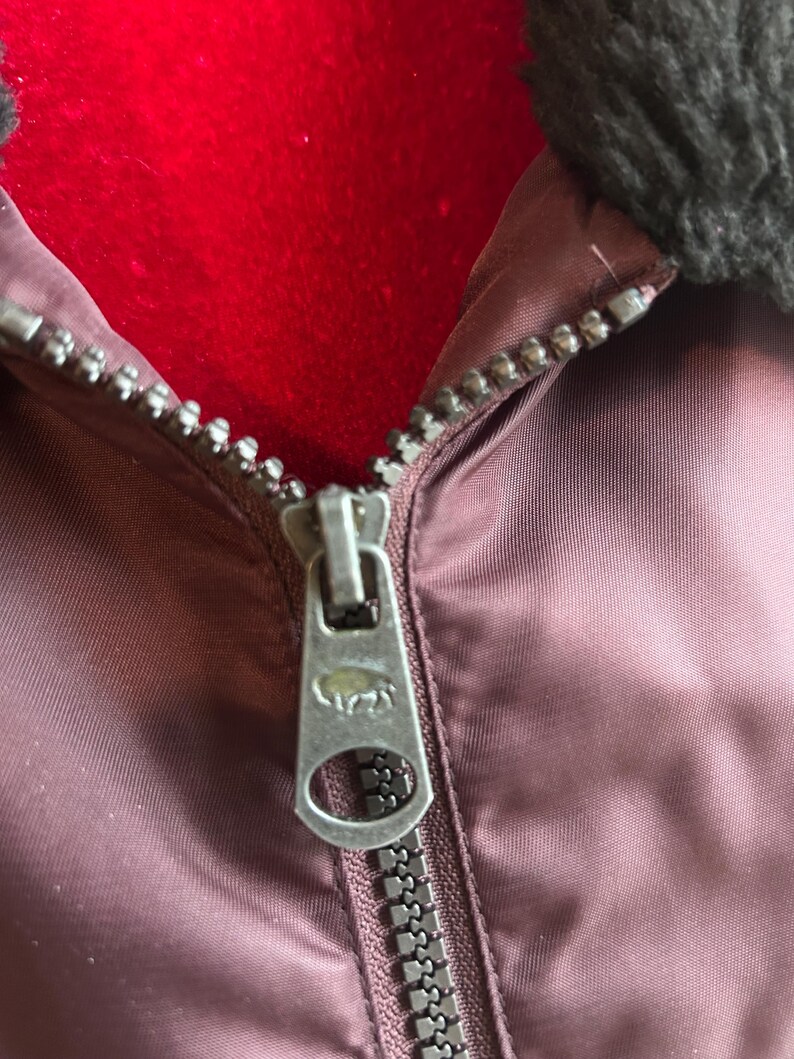 Puede incluir: Primer plano de una cremallera de chaqueta granate. La cremallera plateada tiene un tirador detallado con un dise&ntilde;o de animal. La chaqueta tiene un cuello oscuro y difuso y un interior rojo.