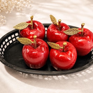 May include: Five glossy red apple figurines with metallic gold stems and textured leaves arranged inside a black oval plastic basket with a lattice-style side design. Each apple features a smooth, reflective surface and a realistic rounded shape.