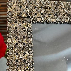May include: An antique-style picture frame with a gold finish. The frame has detailed floral and geometric patterns, with black and clear crystal-like embellishments. The design is intricate and elegant, suitable for displaying photographs or artwork.