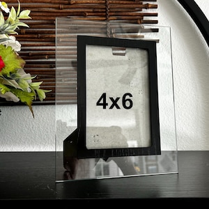 May include: A clear glass photo frame with a black border, designed to hold a 4x6 inch photo. The frame is standing upright on a black surface, with a bamboo blind in the background.