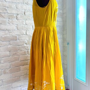 Puede incluir: Un vestido amarillo brillante sin mangas con bordado floral blanco en el dobladillo. El vestido tiene un corpi&ntilde;o ajustado y una falda acampanada. El vestido se exhibe en un maniqu&iacute;. El vestido est&aacute; hecho de una tela ligera.