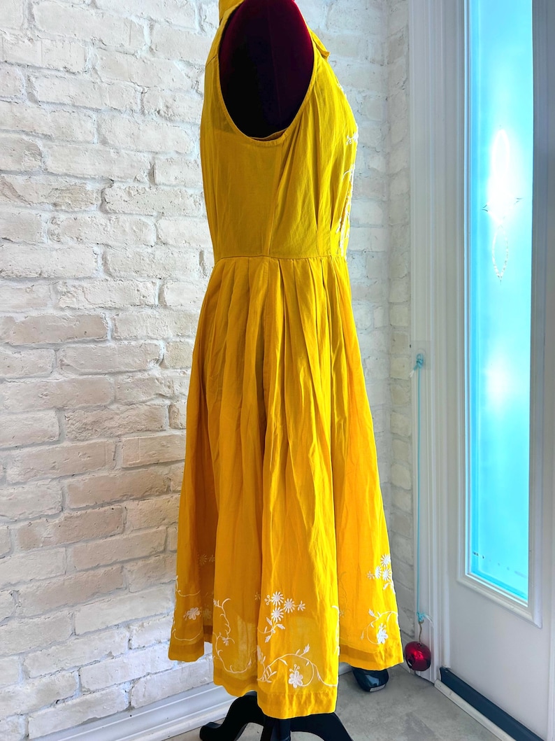 Puede incluir: Un vestido amarillo brillante sin mangas con bordado floral blanco en el dobladillo. El vestido tiene un corpi&ntilde;o ajustado y una falda acampanada. El vestido se exhibe en un maniqu&iacute;. El vestido est&aacute; hecho de una tela ligera.