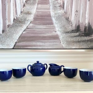 May include: A navy blue tea set with white interiors, including a teapot, creamer, and four teacups. The tea set is arranged on a light-colored surface, with a painting in the background.