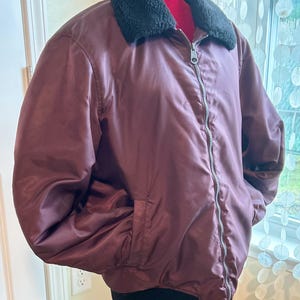 Puede incluir: Chaqueta bomber burdeos con cuello de piel sint&eacute;tica negra y cremallera plateada. La chaqueta tiene dos bolsillos delanteros y un dobladillo acanalado negro. La chaqueta se exhibe en un maniqu&iacute;.