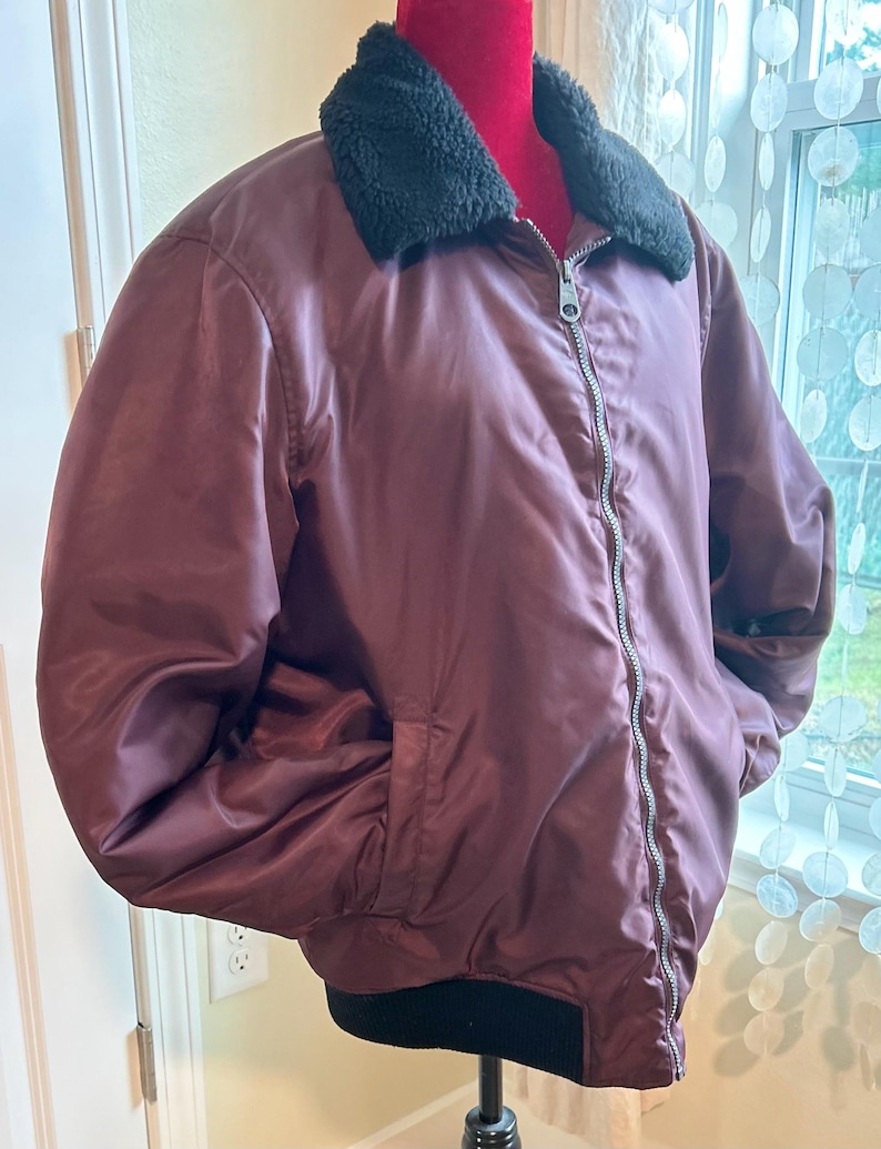 Puede incluir: Chaqueta bomber burdeos con cuello de piel sint&eacute;tica negra y cremallera plateada. La chaqueta tiene dos bolsillos delanteros y un dobladillo acanalado negro. La chaqueta se exhibe en un maniqu&iacute;.