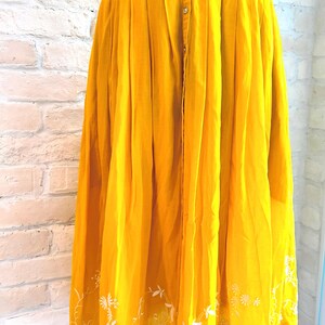 Puede incluir: Vestido amarillo mostaza con bordados florales blancos. El vestido presenta una parte delantera abotonada, una falda plisada y delicados detalles florales bordados a lo largo del escote y el dobladillo. El vestido est&aacute; hecho de tela ligera.