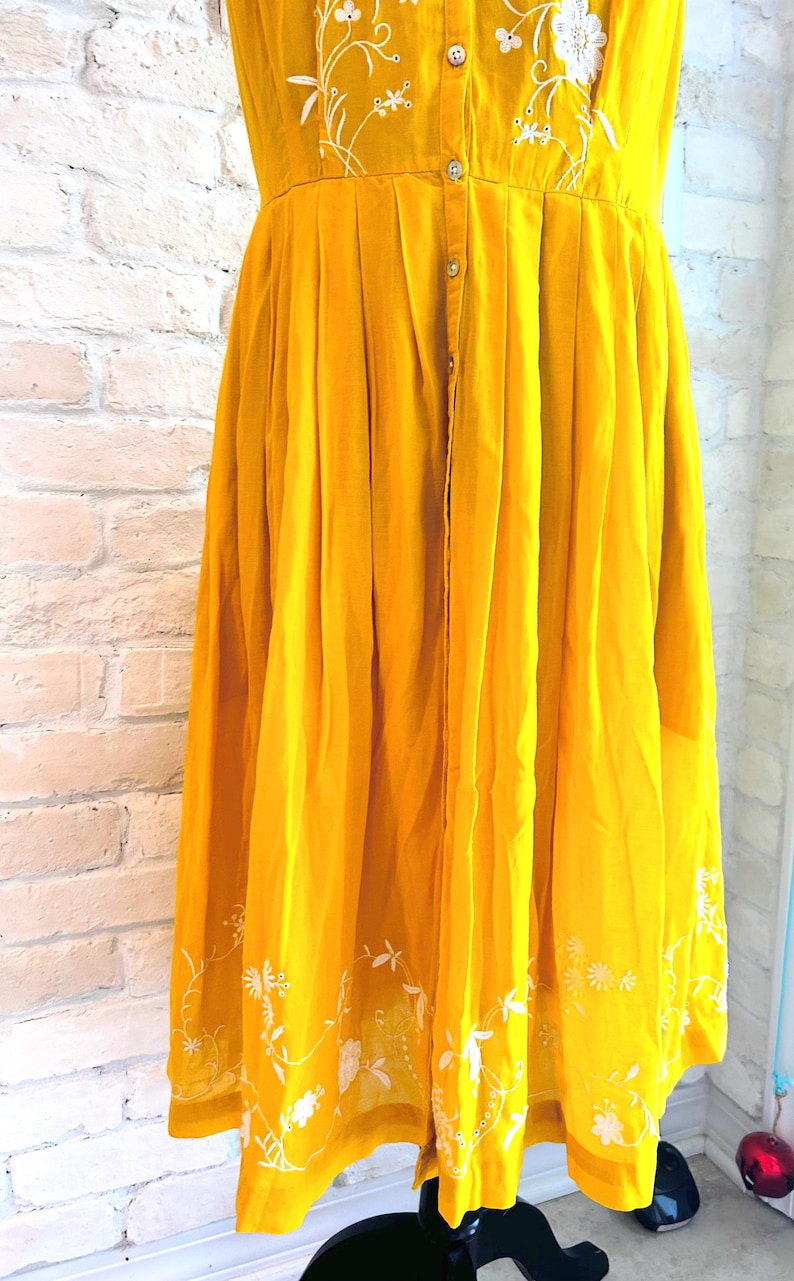 Puede incluir: Vestido amarillo mostaza con bordados florales blancos. El vestido presenta una parte delantera abotonada, una falda plisada y delicados detalles florales bordados a lo largo del escote y el dobladillo. El vestido est&aacute; hecho de tela ligera.