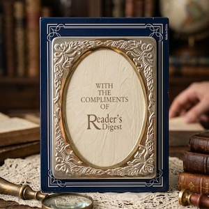 May include: A rectangular dark blue box featuring a central oval silver-toned frame with ornate leaf detailing. The cream-colored insert displays the text "WITH THE COMPLIMENTS OF Reader's Digest" in a dark brown serif font. The box has thin white lines.