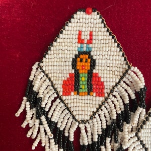 Puede incluir: Pendientes de cuentas blancas con un dise&ntilde;o de cuentas negras y de colores de una mujer nativa americana. Los pendientes tienen una franja de cuentas blancas y negras.
