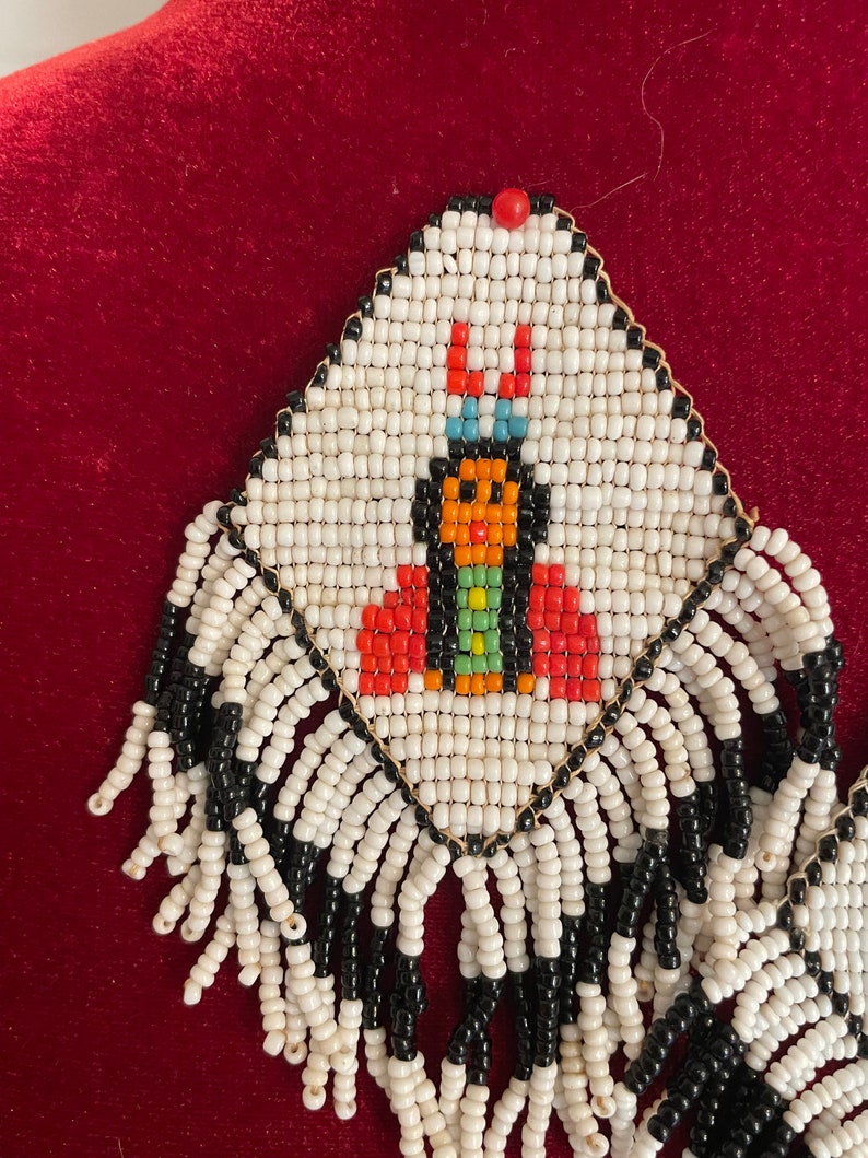 Puede incluir: Pendientes de cuentas blancas con un dise&ntilde;o de cuentas negras y de colores de una mujer nativa americana. Los pendientes tienen una franja de cuentas blancas y negras.