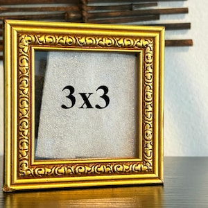 Könnte beinhalten: Ein quadratischer, goldfarbener Bilderrahmen mit aufwendigen Details. Der Rahmen ist mit "3x3" in schwarzem Text beschriftet. Der Rahmen steht auf einer dunklen Oberfläche, mit einem neutralen Hintergrund.