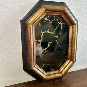 May include: An octagonal wall mirror with a black and gold frame. The mirror's surface is dark green with gold veining. The frame has a gold inner edge and a black outer edge.