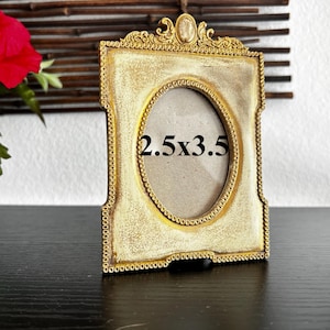 Puede incluir: Marco de fotos dorado de estilo antiguo con abertura ovalada. El marco tiene un acabado envejecido y mide 6,35 x 8,89 cm. El marco tiene detalles decorativos y un pequeño acento ovalado en la parte superior.