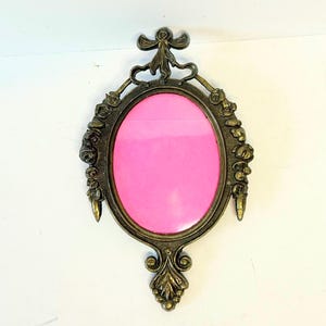 Puede incluir: Un marco de fotos ovalado de metal con un acabado en latón oscuro. El marco presenta detalles ornamentales de flores y cintas a lo largo de los lados y la parte superior, con una base decorativa. El centro contiene una pieza rosa brillante.