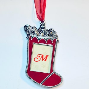 May include: A red and silver-toned metal Christmas stocking ornament featuring a central rectangular photo frame with a red letter M printed on the insert. The top of the stocking is decorated with sculpted toys and a red ribbon loop for hanging.
