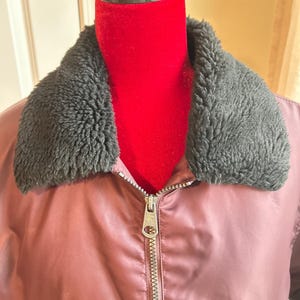 Puede incluir: Chaqueta bomber granate con cuello de piel sint&eacute;tica negra. La chaqueta tiene cierre de cremallera plateada y un acabado satinado. La chaqueta se exhibe en un maniqu&iacute; rojo.