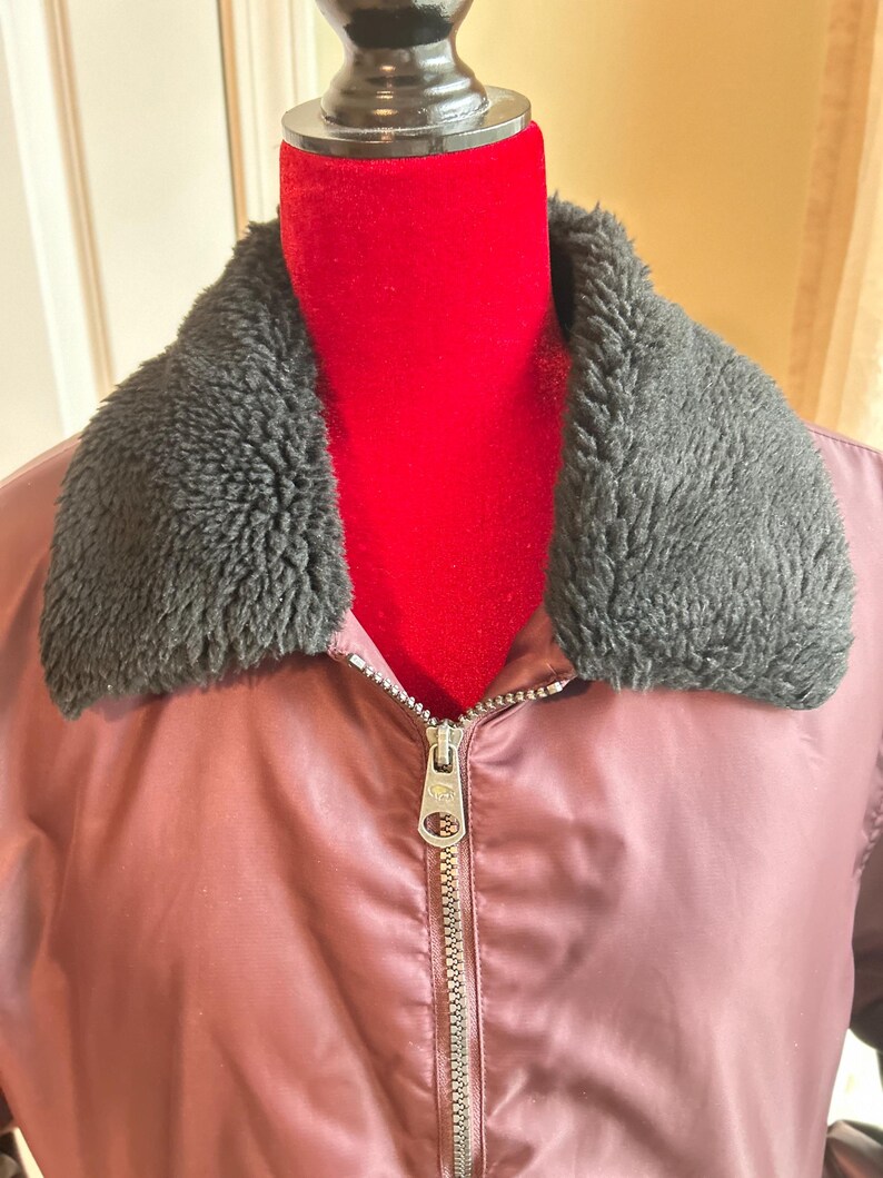 Puede incluir: Chaqueta bomber granate con cuello de piel sint&eacute;tica negra. La chaqueta tiene cierre de cremallera plateada y un acabado satinado. La chaqueta se exhibe en un maniqu&iacute; rojo.