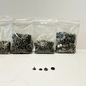 May include: Four clear plastic bags filled with various sizes of metal snap fasteners. The snaps are in shades of silver and gunmetal. Several individual snaps are arranged below the bags.