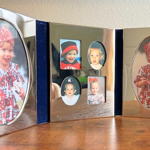 May include: A silver-toned, tri-fold picture frame with multiple oval and rectangular openings. The frame holds several photographs of children. The frame is displayed on a wooden surface.