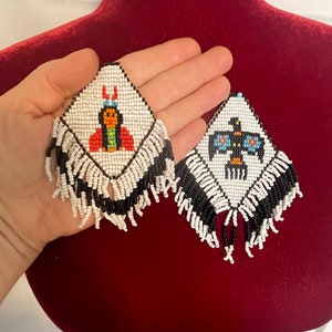 Puede incluir: Un par de pendientes de cuentas blancas y negras con flecos. Un pendiente presenta una imagen de cuentas de una mujer nativa americana con ropa tradicional, mientras que el otro pendiente presenta una imagen de cuentas de un p&aacute;jaro con las alas extendidas.